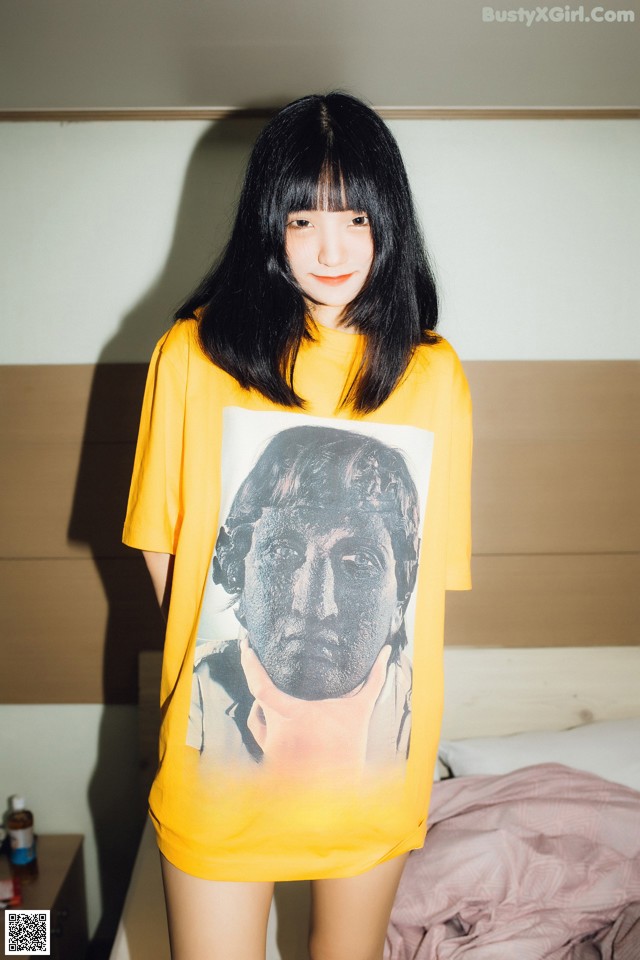 A woman standing in front of a bed wearing a yellow shirt with a picture of Paul McCartney on it.